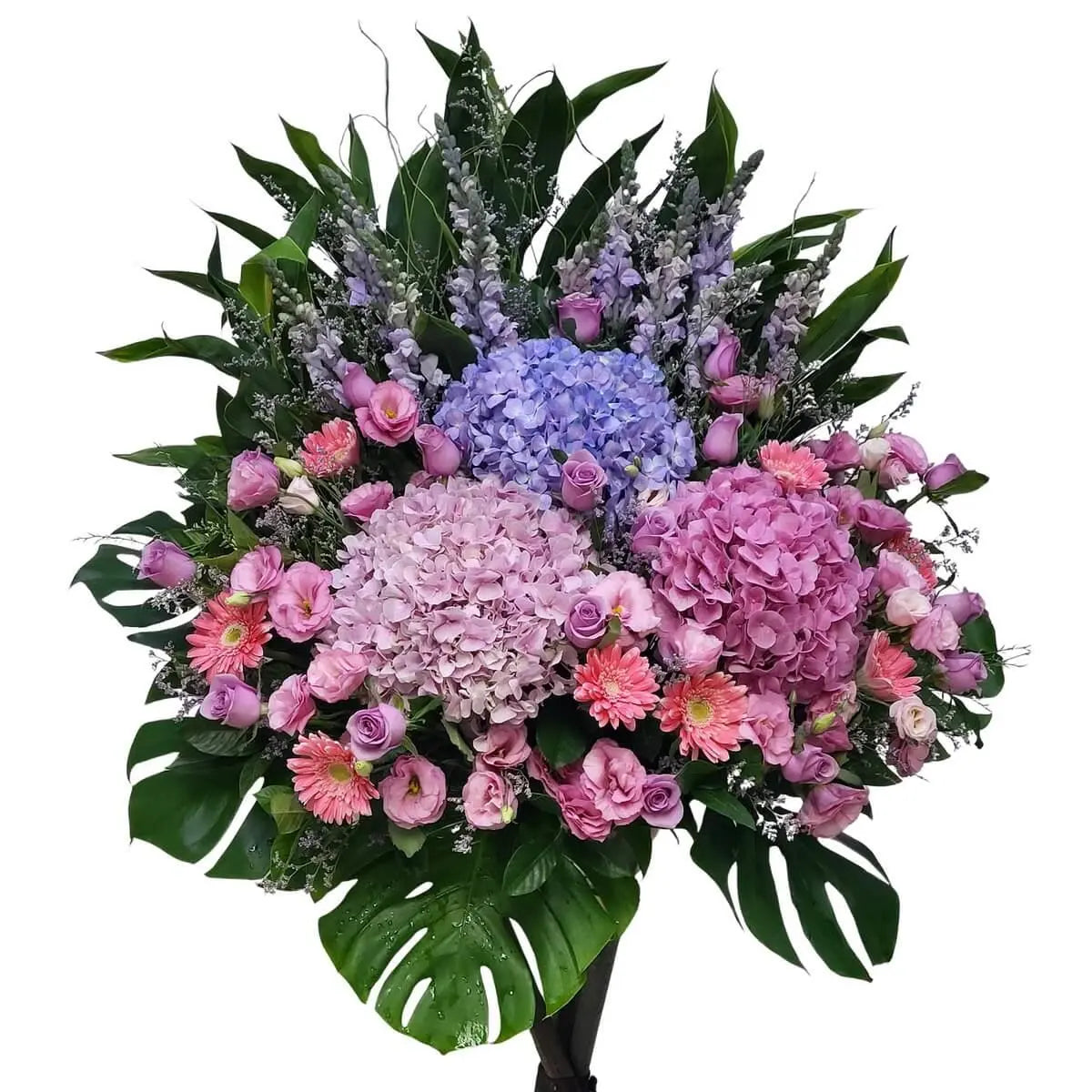 Large standing arrangement of pink and blue hydrangeas, pink gerberas, lisianthus, snapdragons, and lavender roses with tropical greenery | 粉藍色繡球花配搭粉紅非洲菊、洋桔梗、金魚草及紫玫瑰,襯以熱帶綠葉製成大型站立式花牌。