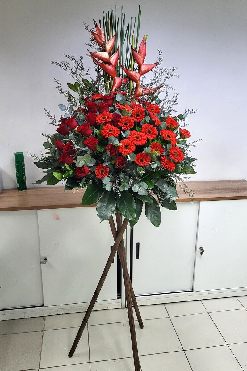 Original vertical flower stand with red heliconias, red roses, and red gerberas, arranged with eucalyptus and lush greenery on a wooden tripod base, displayed indoors - 紅赫蕉、紅玫瑰與紅扶郎花牌,配尤加利與繁葉,木三腳架承托,室內陳列