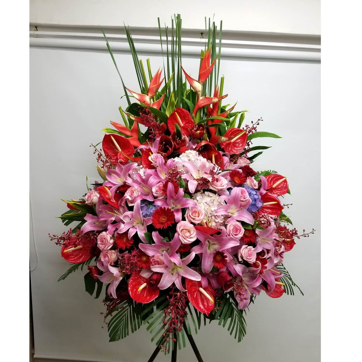 Striking floral stand featuring pink lilies, red anthuriums, cymbidium orchids, pink roses, red gerberas, hydrangeas, and heliconia.
艷麗花牌以粉百合、紅火鶴、蘭花、玫瑰、非洲菊與繡球花組成,層次豐富,襯以熱帶綠葉。