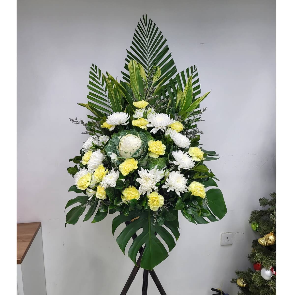 Elegant condolence arrangement of white chrysanthemums, yellow carnations, and ornamental cabbage, with layered tropical foliage. 白菊、黃康乃馨與觀賞包菜配以綠葉層次,打造莊重哀悼花牌。
