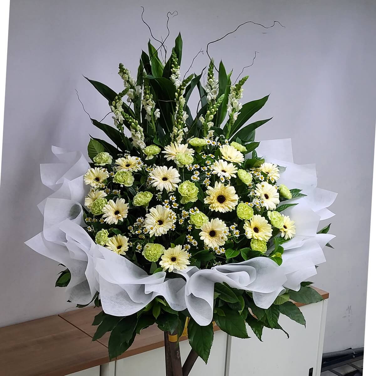 Elegant condolence stand with white gerberas, light green carnations, chamomile, and lush foliage, softened by a white fabric wrap – 優雅悼念花籃,白太陽菊、淡綠康乃馨與洋甘菊搭配綠葉與白紗,莊重素雅