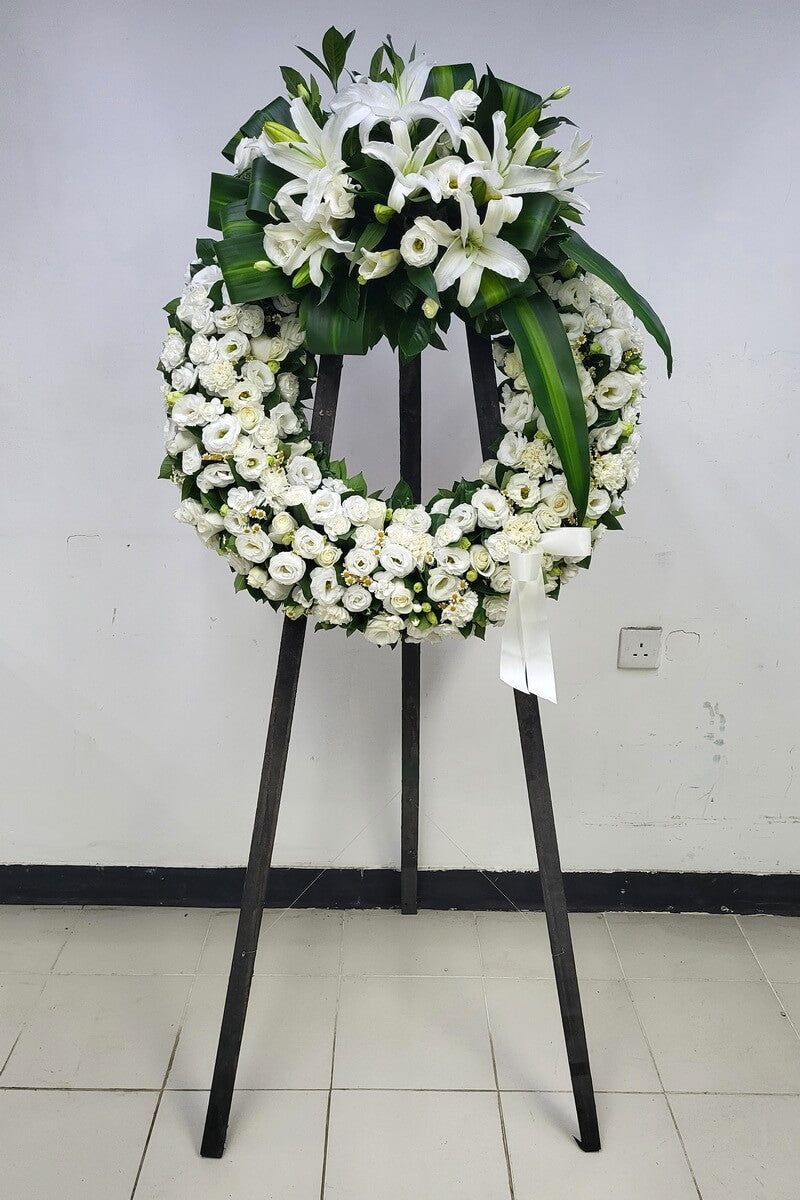 Full floral wreath featuring white lilies, roses, lisianthus, carnations, chamomile daisies, structured tropical greenery and a ribbon, presented on black easel stand - 白百合、玫瑰、洋桔梗、康乃馨、黃芯小白菊及熱帶綠葉配絲帶花圈擺放於黑色支架上