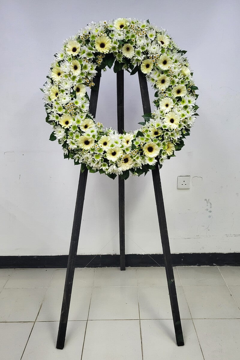 Round floral wreath featuring white-yellow gerberas, spray and disbud chrysanthemums with green centers, chamomile-like daisies and green leaves, displayed on a black tripod - 白黃色太陽菊、噴菊、綠芯大頭菊、小白菊及綠葉組合圓形花圈展示於黑色三腳支架上