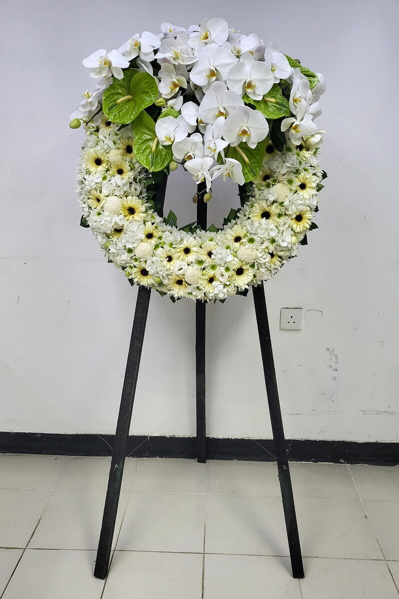 Full view of standing floral wreath with cascading white phalaenopsis orchids and green anthuriums above a base of cream gerberas, button chrysanthemums, and clustered white hydrangeas 白色蝴蝶蘭與綠色火鶴花垂墜設計,搭配奶白色非洲菊、乒乓菊及繡球花的完整立式花圈圖