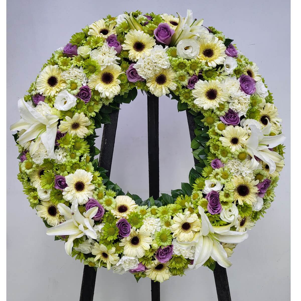 Full circular wreath composed of white gerbera daisies, white lilies, white lisianthus, green button chrysanthemums, and purple roses, arranged with dense layering and balanced green foliage 白色非洲菊、白色百合、白色洋桔梗、綠色菊花及紫色玫瑰花圈設計