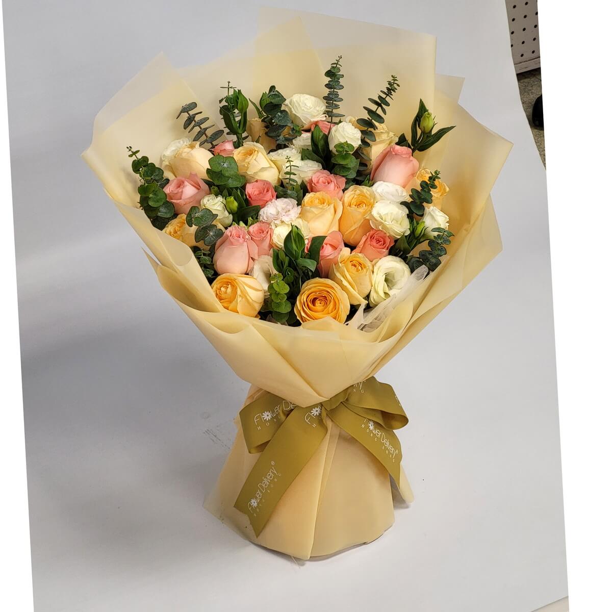 Hand-tied bouquet of peach, cream, and blush roses with eucalyptus and foliage 淡橙、奶白及粉紅玫瑰組合搭配尤加利葉與綠葉,手綁花束以柔黃包裝紙及金絲帶包裹,色調溫柔自然。