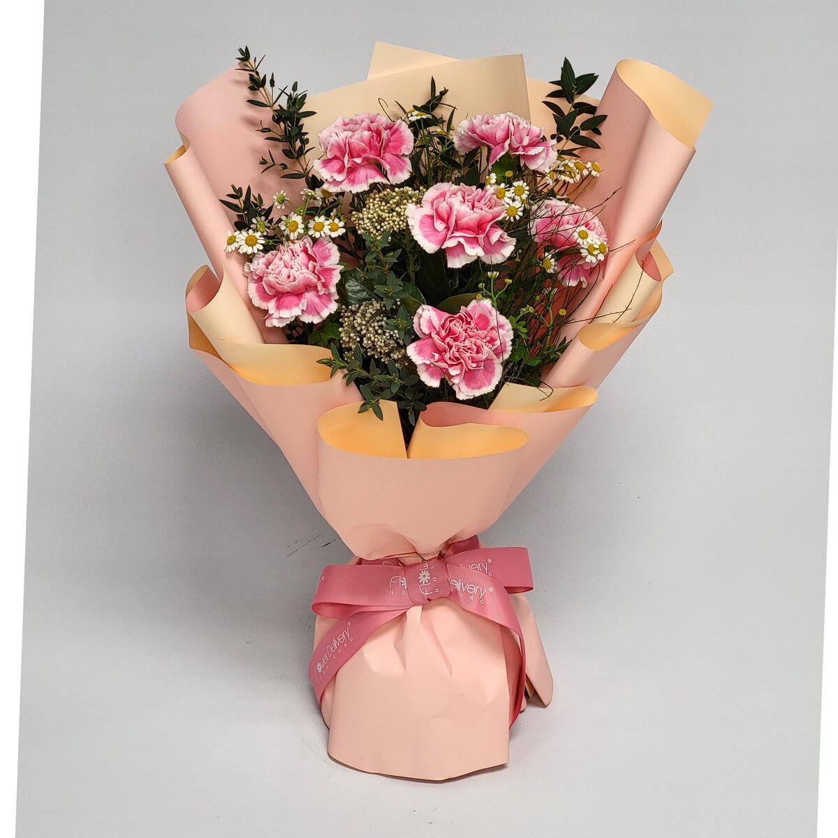Hand-tied bouquet of pink bi-color carnations, chamomile, and white yarrow wrapped in peach-toned paper 雙色粉紅康乃馨、洋甘菊及白蓍草搭配綠葉,蜜桃橙色包裝紙及粉紅絲帶束口設計。