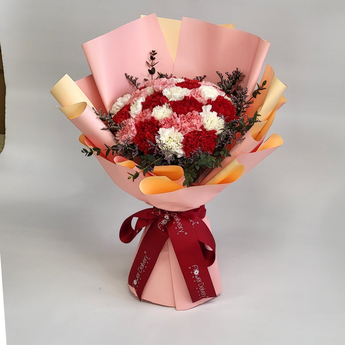 Hand-tied bouquet of red, white, and pink carnations with foliage, wrapped in layered orange-pink paper 紅白粉康乃馨搭配葉材製成手綁花束,橙粉色層次包裝紙及酒紅色絲帶束口。