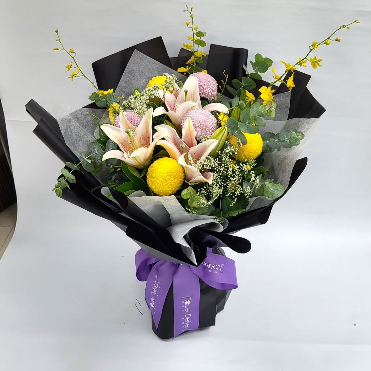 Arrangement of pink lilies, yellow and pink button chrysanthemums, yellow oncidium, baby's breath, and eucalyptus in black-grey wrap with purple ribbon. 粉紅百合、粉黃乒乓菊、文心蘭、滿天星及尤加利葉花束，黑灰色包裝，紫色絲帶。