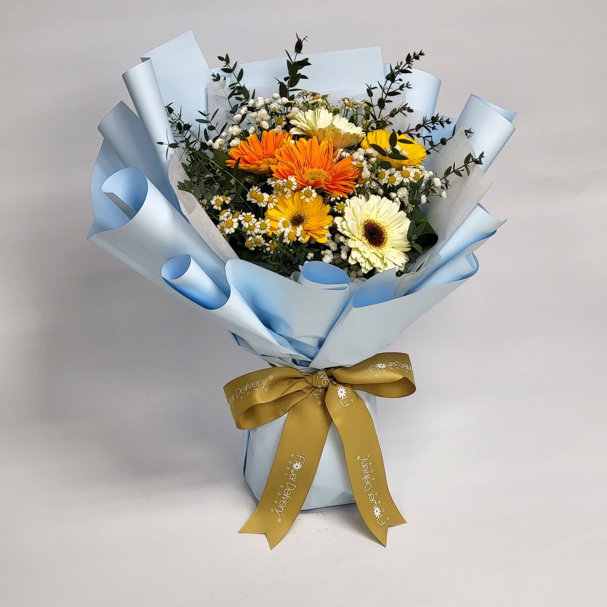 Arrangement of orange, yellow, and cream gerbera daisies with chamomile, baby’s breath, and greenery, wrapped in soft blue paper and tied with gold ribbon. 橙、黃、奶白太陽菊配洋甘菊、滿天星及綠葉，淺藍包裝，金色絲帶裝飾。