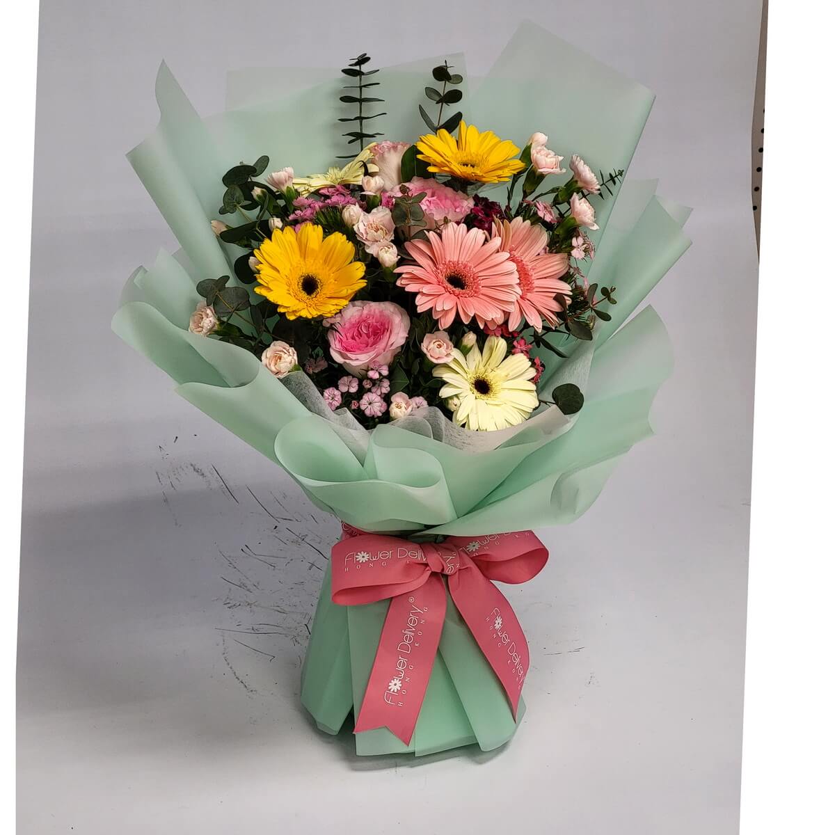 Arrangement of pink, yellow, and cream gerbera daisies, spray roses, and eucalyptus, wrapped in mint green paper and tied with a pink ribbon. 粉紅、黃色及奶白太陽菊、迷你玫瑰和尤加利葉花束，薄荷綠包裝，配粉紅絲帶。