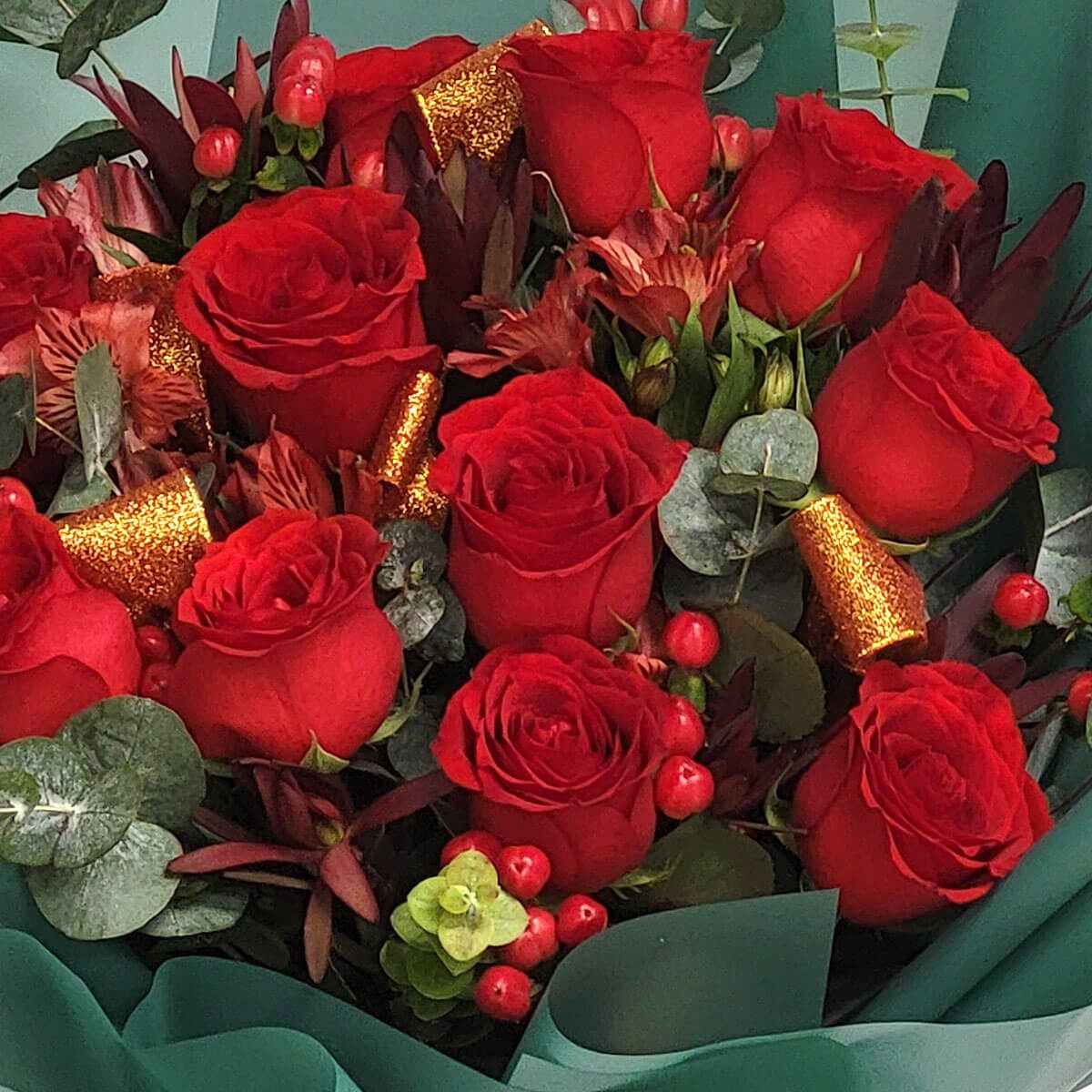 Arrangement of red roses, hypericum berries, eucalyptus, and gold glitter accents 紅玫瑰花束，配以紅色鼠尾草果、尤加利葉與深色葉材，點綴金色亮片，色彩飽滿鮮明。