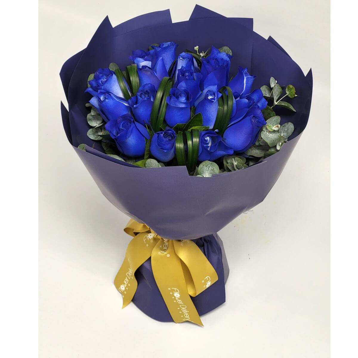Arrangement of vivid blue roses with eucalyptus and green accents, wrapped in deep indigo paper and finished with a gold ribbon. 鮮藍玫瑰花束配尤加利葉及綠葉點綴，深靛色包裝，綁上金色絲帶。