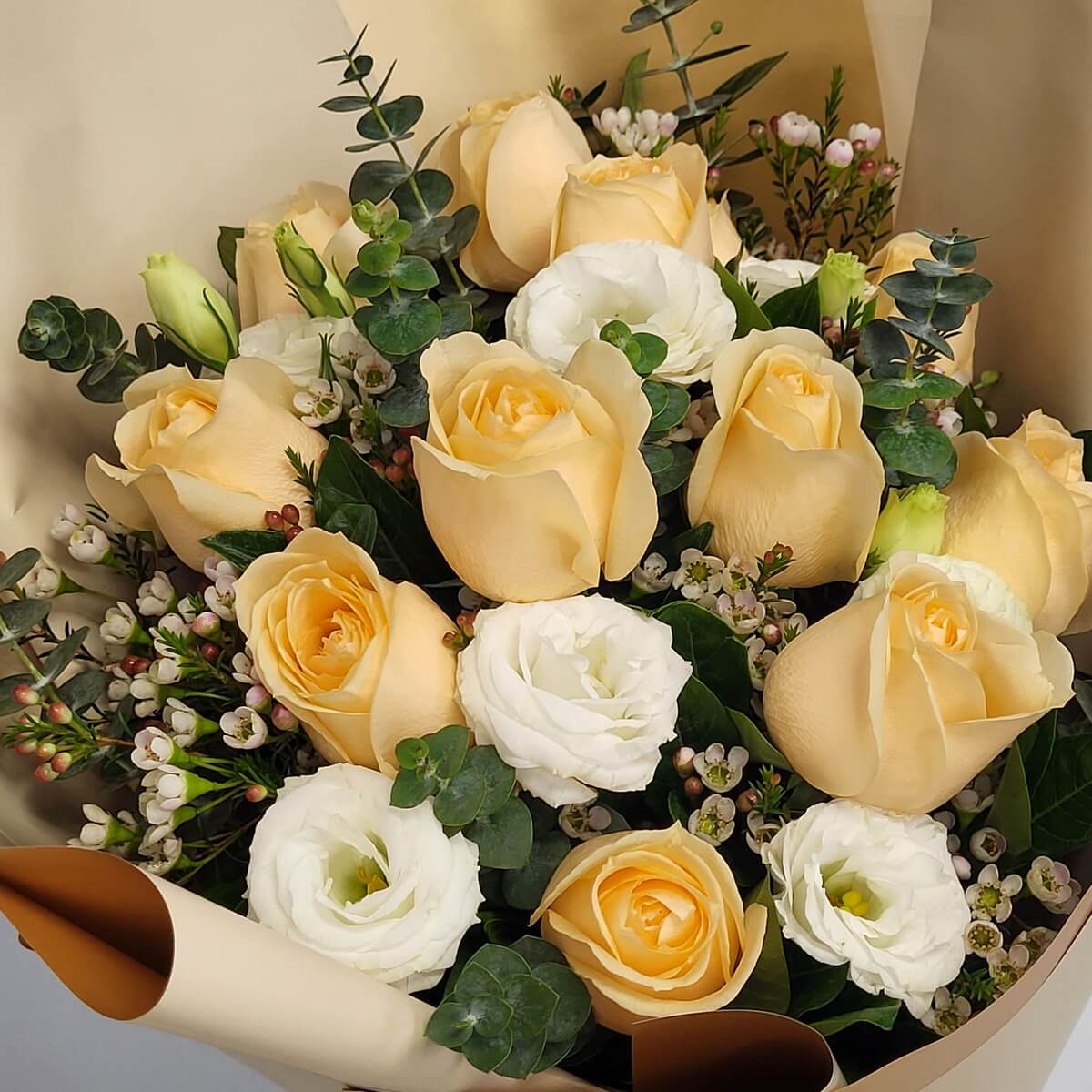 Detail view of champagne-colored roses mixed with white lisianthus, delicate pink wax flowers, and lush eucalyptus foliage in cream wrapping - 香檳玫瑰配白洋桔梗、粉色蠟花及茂密尤加利葉，米白包裝中特寫