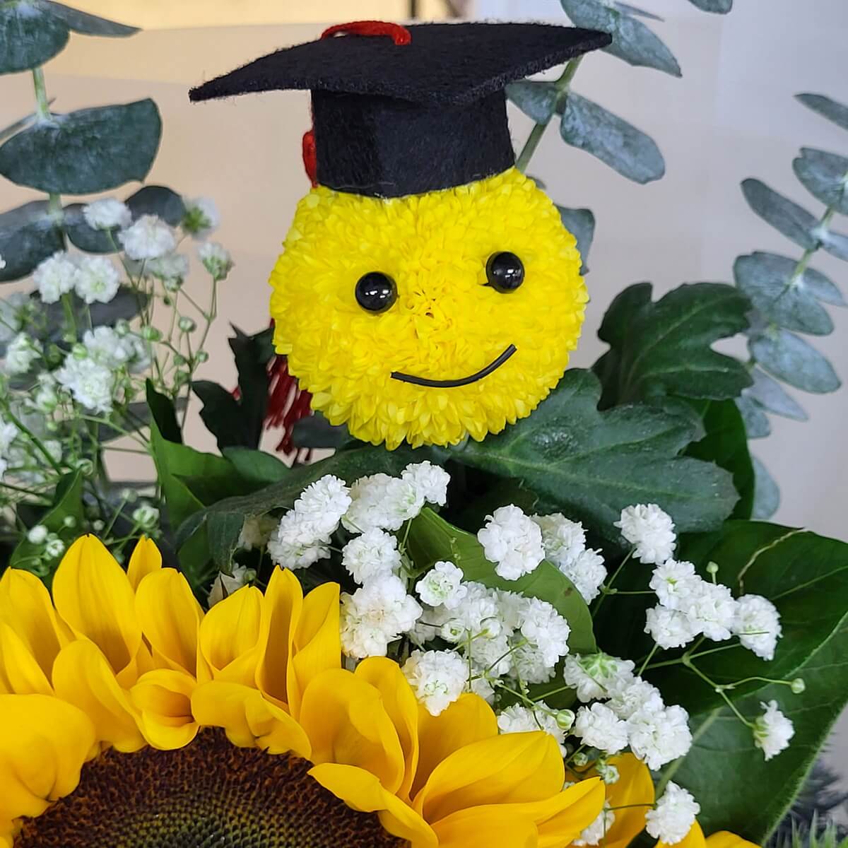 Detail view of graduation bouquet with sunflower, baby’s breath, eucalyptus leaves, and yellow button chrysanthemum smiley face wearing mortarboard 向日葵、滿天星、尤加利葉與戴畢業帽的黃色乒乓菊笑臉造型組成的畢業花束特寫