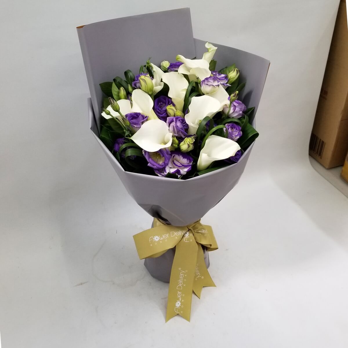 A refined bouquet of white calla lilies and purple lisianthus wrapped in a sleek grey wrapper with a gold ribbon. 高雅白色馬蹄蓮搭配神秘紫色洋桔梗，適合表達尊重、懷念或感謝。