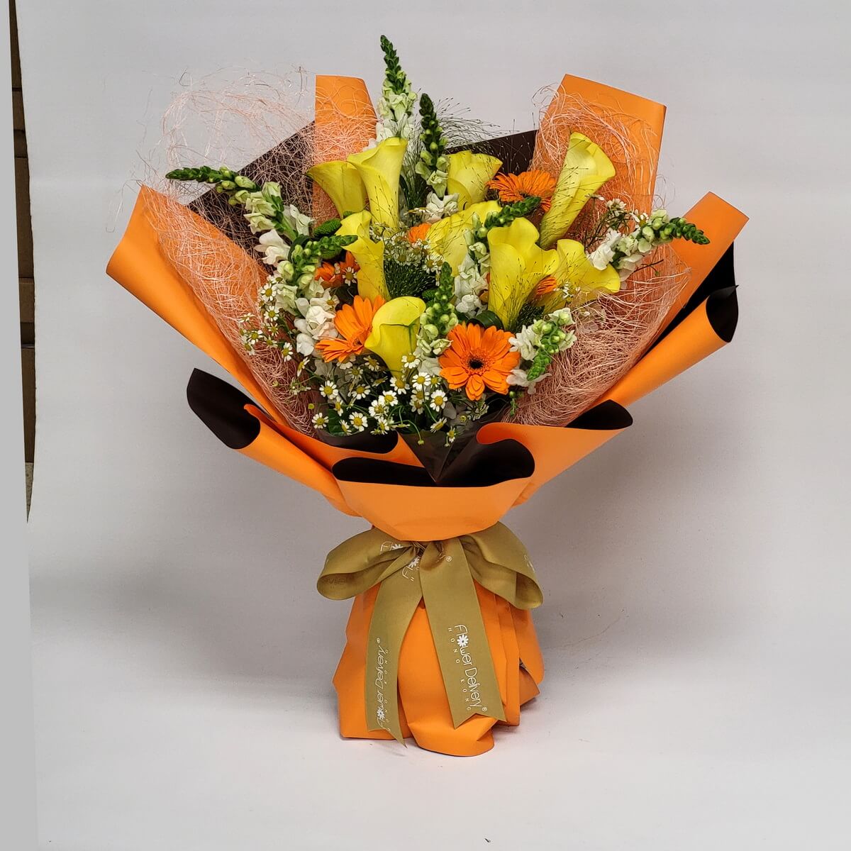 A lively bouquet featuring yellow calla lilies, orange gerberas, white snapdragons, chamomile, and textured accents, wrapped in bright orange and brown paper with a golden ribbon. 熾熱暖陽色系花束，象徵活力與喜悅。