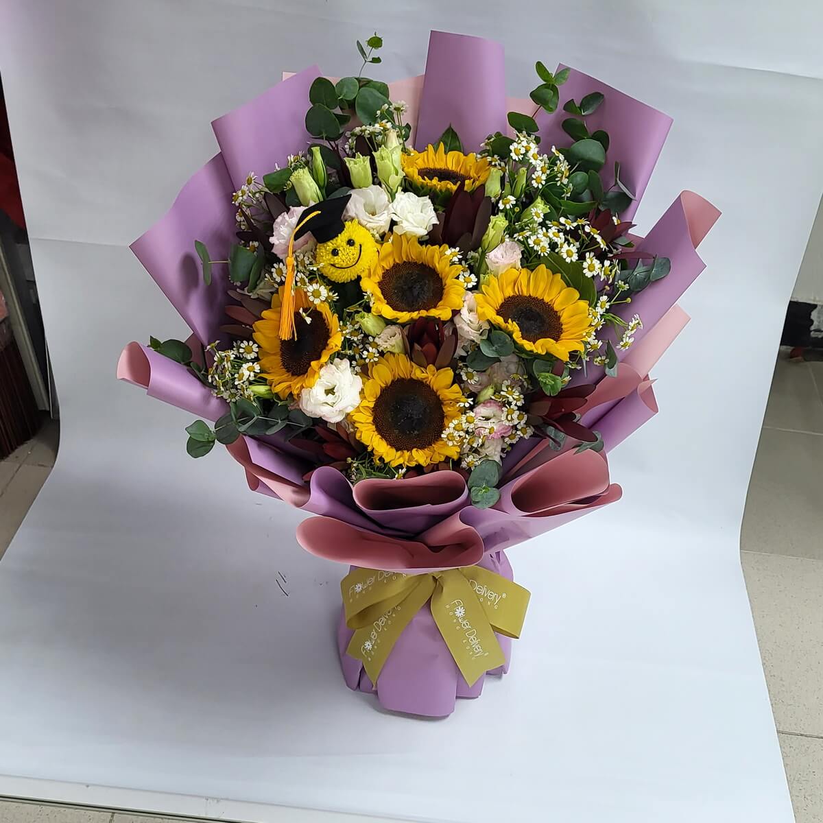 Vibrant bouquet of sunflowers, lisianthus, and eucalyptus, featuring a smiling graduation plush, wrapped in lavender paper with a gold ribbon. 向日葵畢業花束，象徵光明未來與祝福，包裝典雅溫馨。
