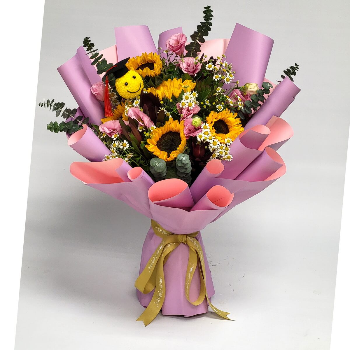 Graduation-themed bouquet wrapped in pink paper, featuring sunflowers, chamomile daisies, pink lisianthus, eucalyptus, and yellow smiley button chrysanthemum 戴畢業帽黃色笑臉乒乓菊、向日葵、洋甘菊、粉洋桔梗與尤加利葉，包裝於粉色紙中的畢業花束