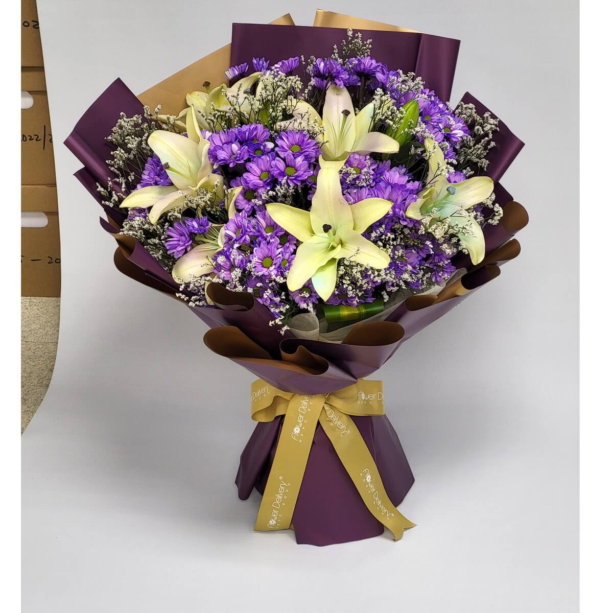 A striking bouquet of creamy lilies and vibrant violet daisies, wrapped in deep plum and gold paper with matching ribbon. 月光下的優雅，紫白花卉交織出浪漫樂章。