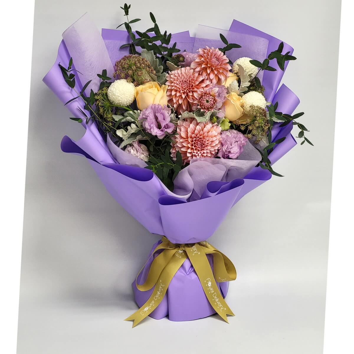 A romantic bouquet with blush dahlias, soft yellow roses, white pompom mums, and lavender lisianthus, surrounded by lush greens and wrapped in lavender paper with a gold ribbon. 柔和配色，散發夢幻與優雅氣息。