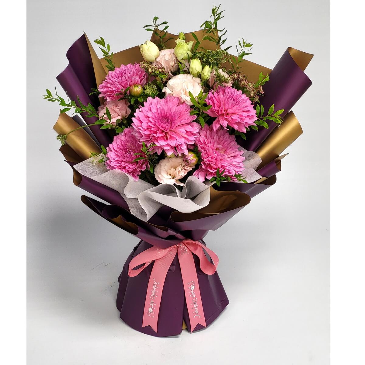 A bouquet featuring bold pink chrysanthemums, blush roses, and buds, wrapped in plum and gold-toned paper with a soft pink ribbon. 鮮明粉菊搭配裸粉玫瑰與花苞，外層是酒紅與金色包裝紙，綁以粉紅緞帶，充滿甜美與尊貴感。