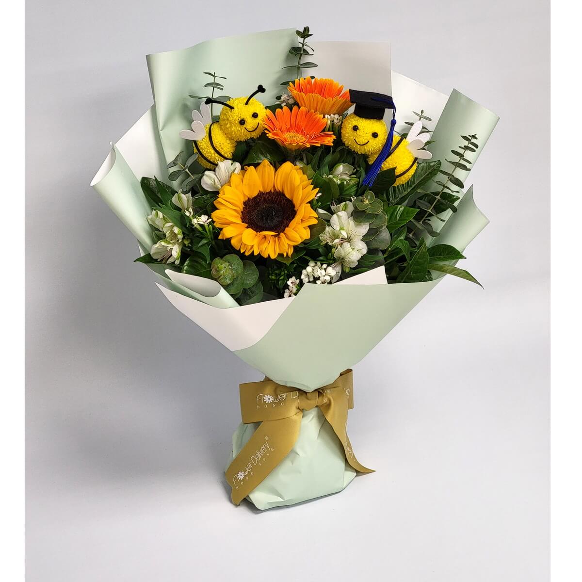 Graduation bouquet with a bright sunflower, orange gerberas, white blooms, eucalyptus, and two yellow pompom bees—one wearing a graduation cap. 包含向日葵、非洲菊、白花與畢業帽毛球蜜蜂，展現青春活力與慶賀氛圍。