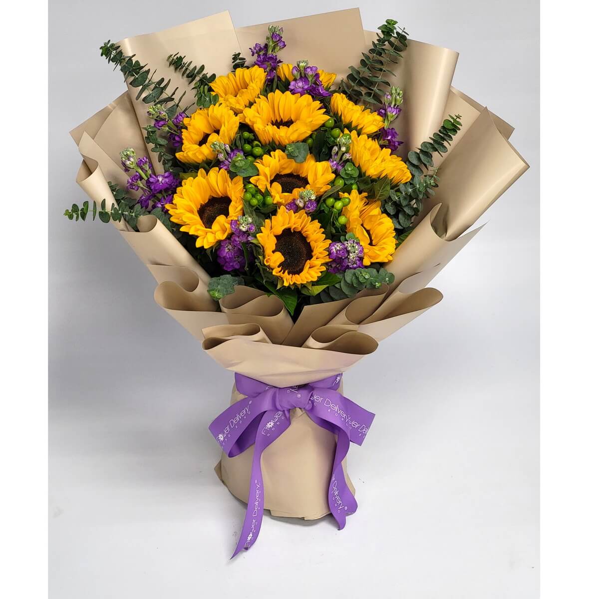 A bright sunflower bouquet accented with vivid purple statice and eucalyptus, wrapped in champagne-colored paper with a purple ribbon. 向日葵與紫色花材交織，搭配尤加利葉與香檳色包裝，展現熱情與優雅。