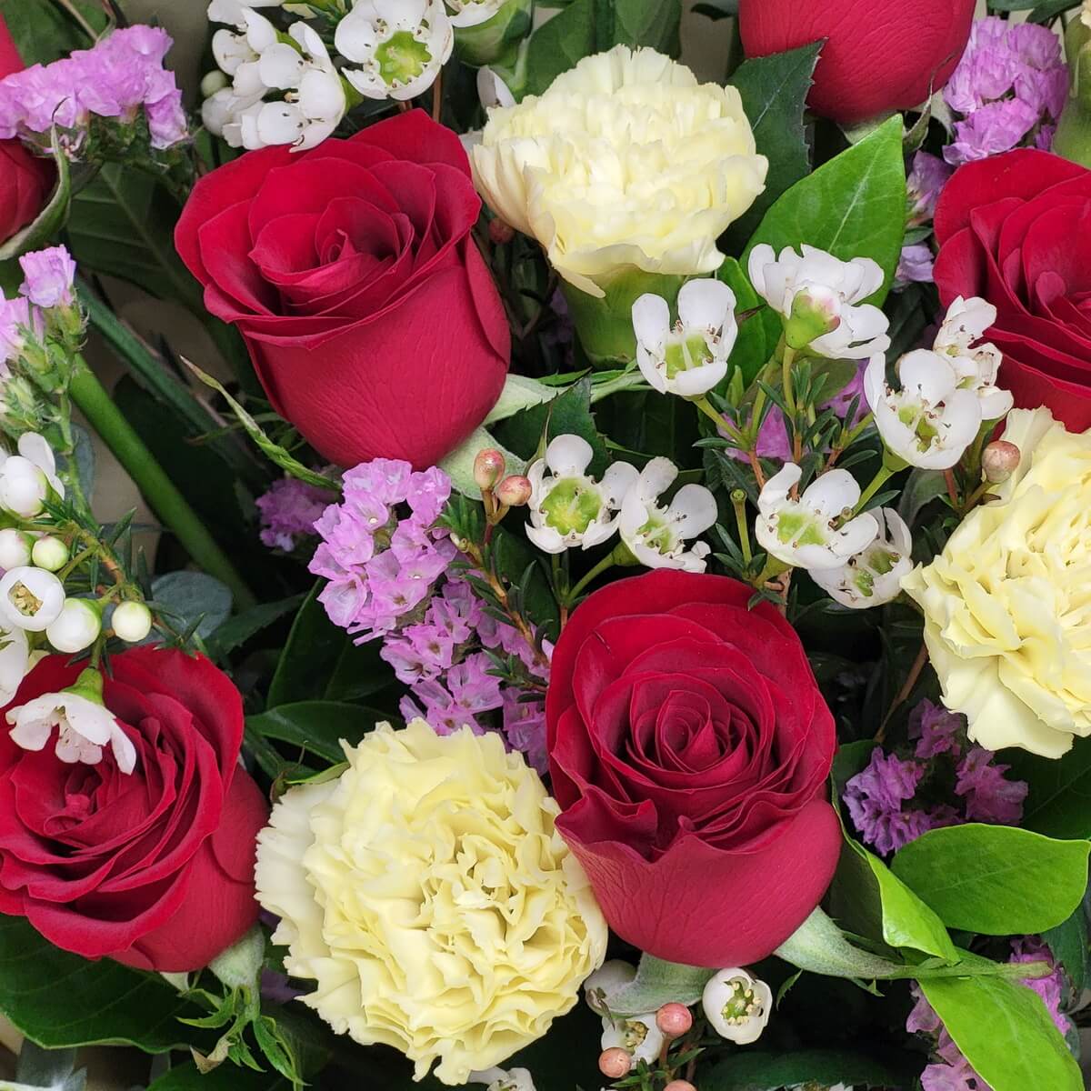 Top view of red roses, soft yellow carnations, white waxflowers, and purple statice flowers with lush green foliage. 紅玫瑰、柔黃康乃馨、白色蠟花與紫色勿忘草混合綠葉的俯視花材。