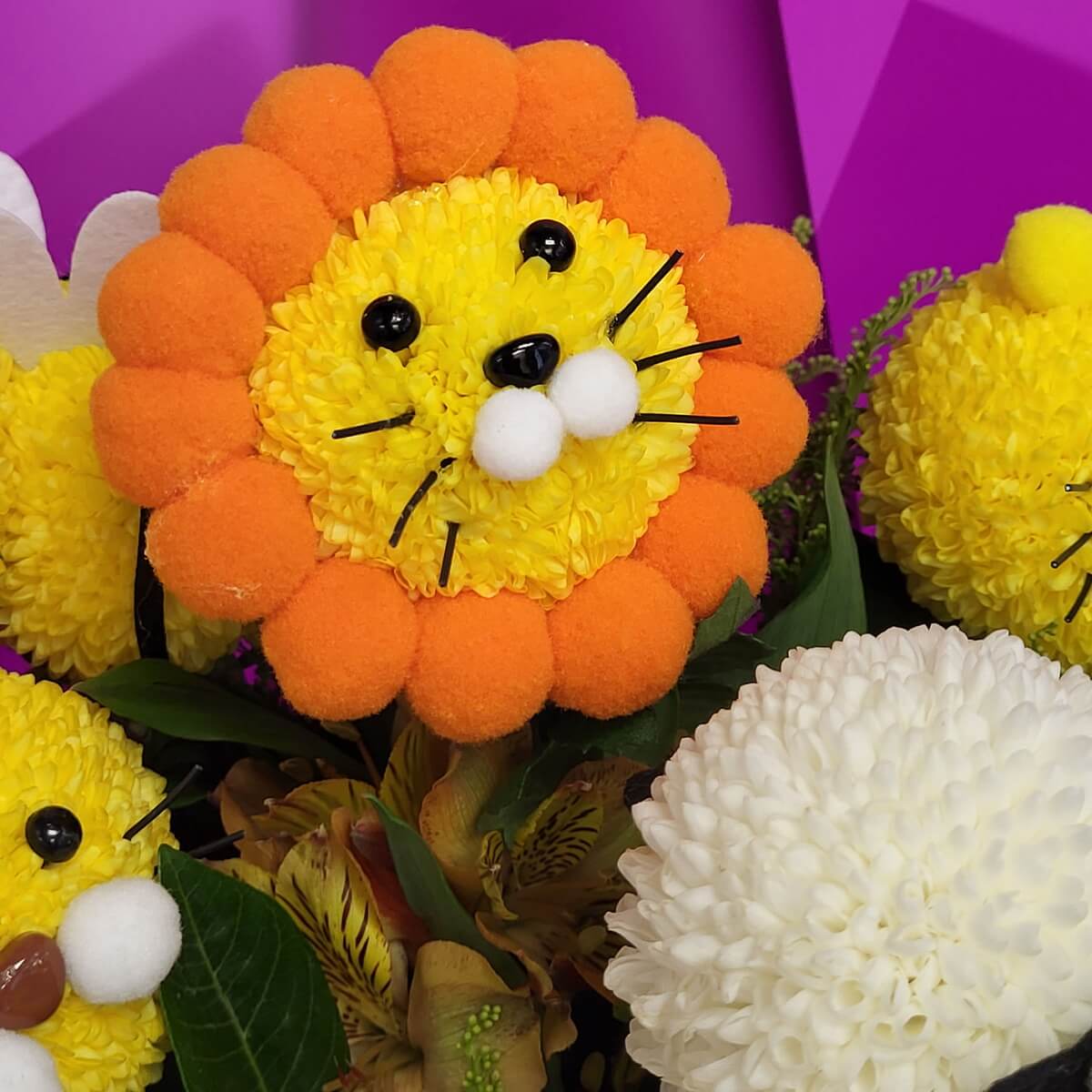 Detail view of a lion head made from yellow button chrysanthemums, accented with black beads for eyes, white pom-pom snout, and orange pom-pom mane - 黃乒乓菊製獅子頭，配黑珠眼、白毛球鼻及橙毛球鬃毛特寫