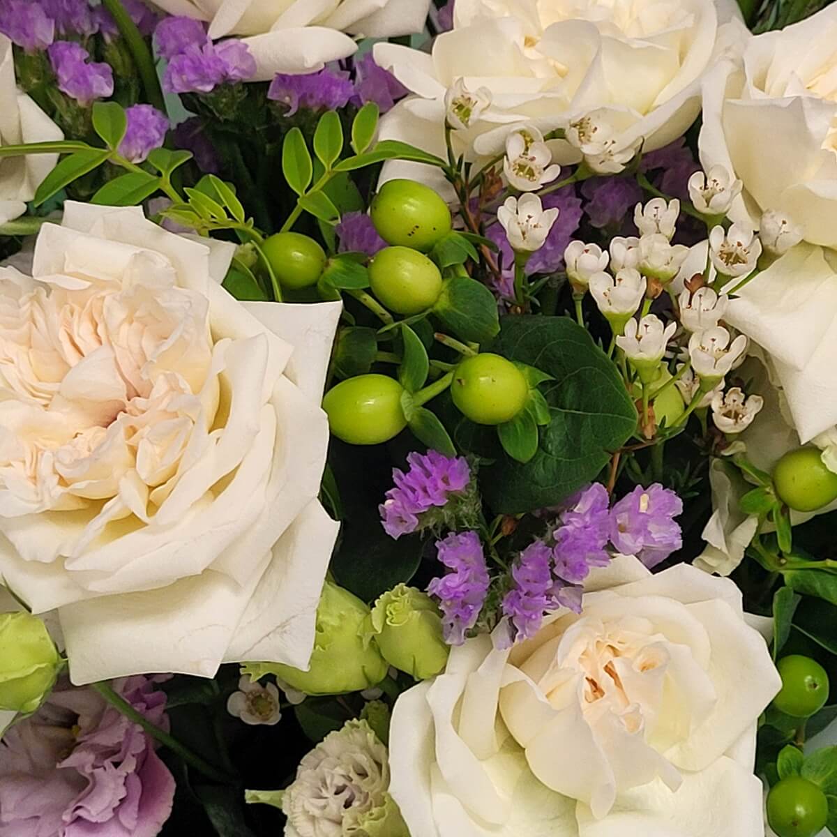 Top view of champagne roses, accented by white waxflowers, green hypericum berries, and lavender statice with rich foliage. 香檳色玫瑰配白色蠟花、綠色貝母果與淡紫勿忘草的混合花材俯視圖。