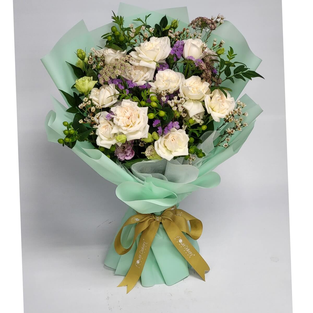 Delicate bouquet of ivory roses, purple statice, wax flowers, and greenery, wrapped in mint green paper with a gold ribbon. 粉白玫瑰與紫色花材點綴，搭配綠色包裝，展現優雅氣質。