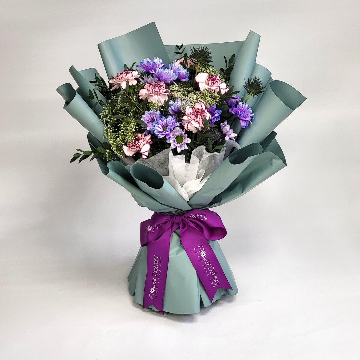 A striking bouquet featuring lavender and purple daisies, two-tone pink carnations, Queen Anne’s lace, and blue eryngium thistle. Wrapped in muted teal paper and tied with a rich purple satin ribbon. 夢幻清新，極具現代感。