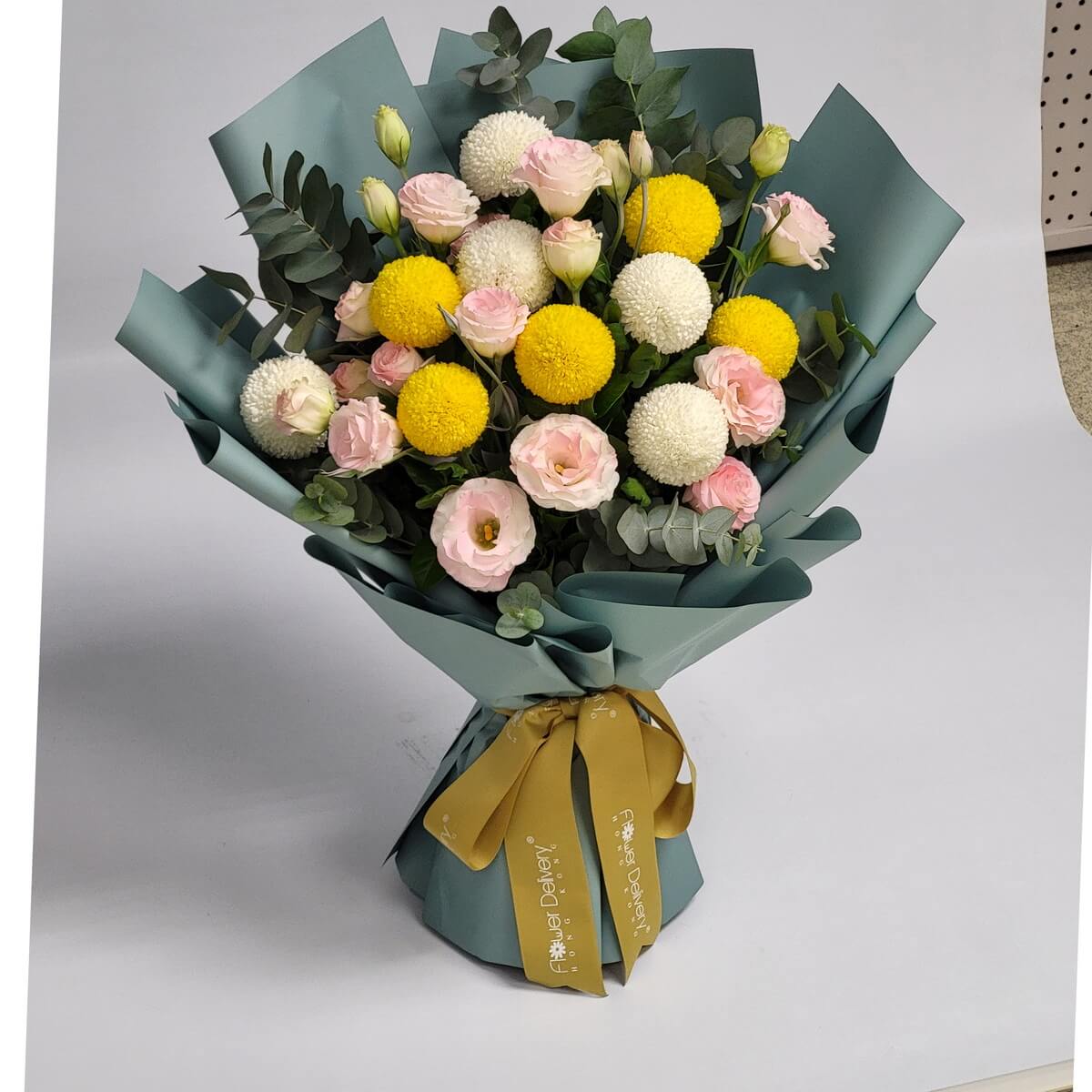 A lively bouquet featuring yellow and white billy balls (craspedia and ping pong mums), soft pink eustoma, and buds, arranged with eucalyptus foliage and wrapped in matte teal paper with a gold ribbon. 活潑明亮，洋溢著溫柔與希望。