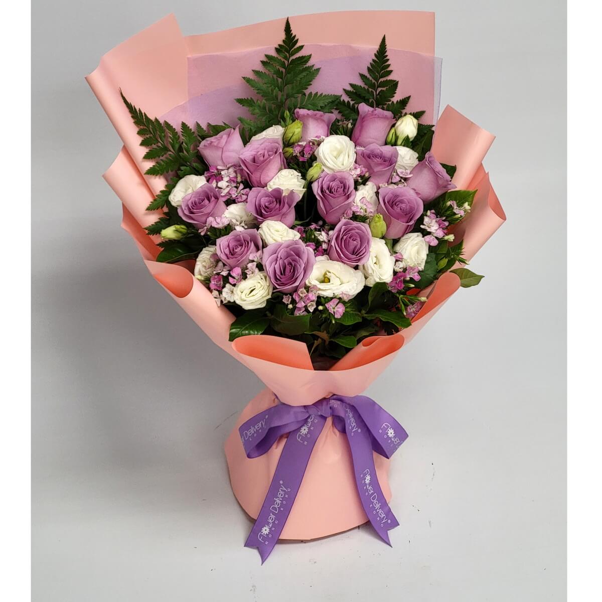 An enchanting bouquet of lavender and white roses, accented with dainty pink fillers and fern leaves, wrapped in peach and lilac paper with a purple ribbon. 紫白玫瑰交織，優雅脫俗，散發夢幻般的浪漫氣息。