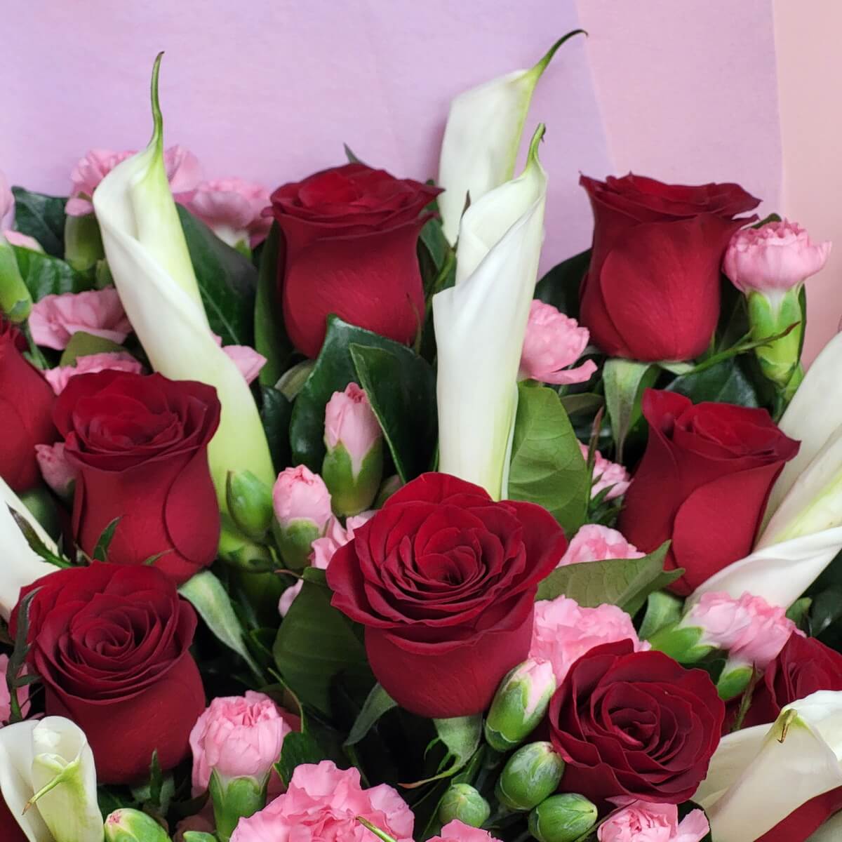 Detailed close-up of red roses, white calla lilies, and pink spray carnations arranged among lush green leaves 紅玫瑰、白馬蹄蘭與粉紅迷你康乃馨，配搭濃密綠葉的細節特寫