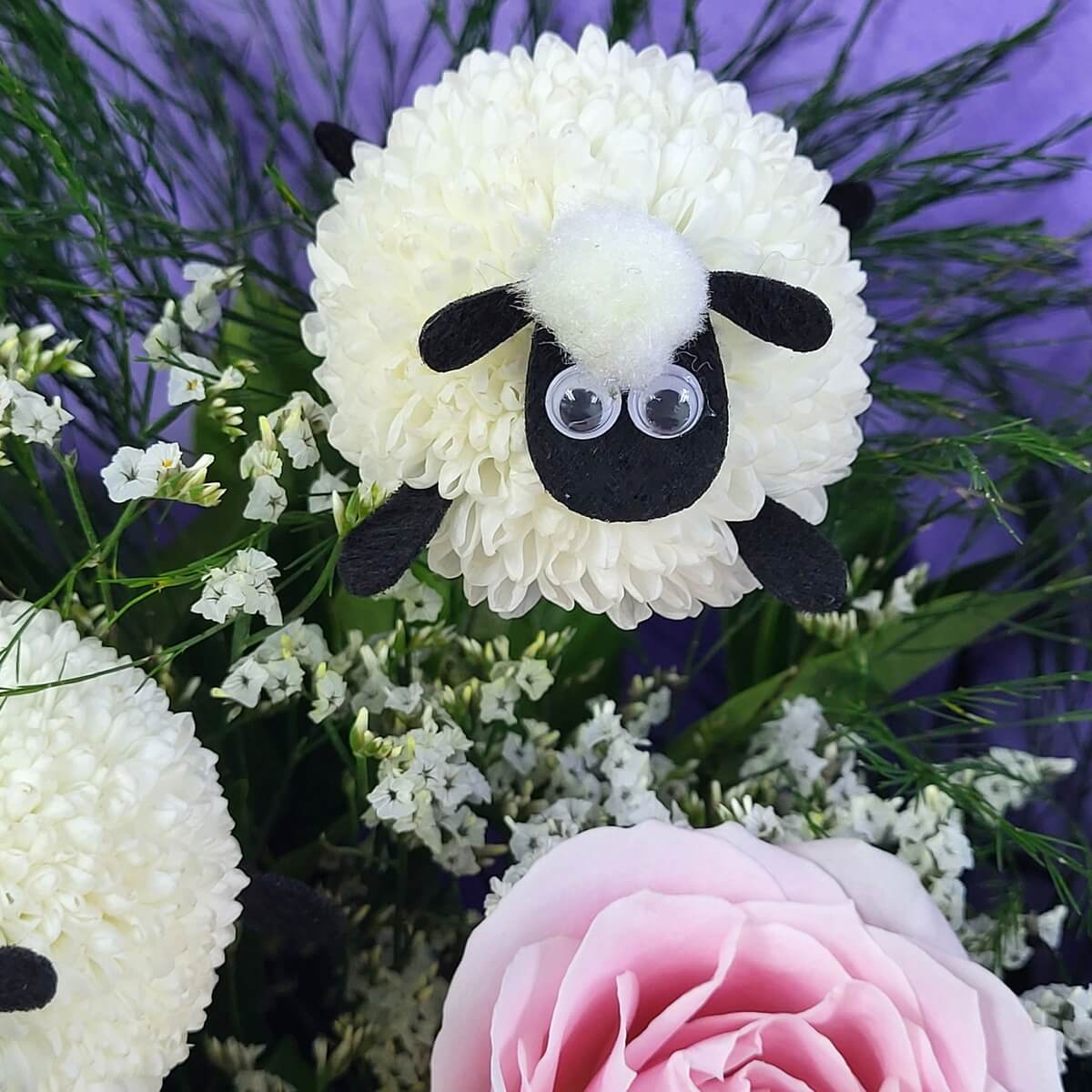 Close-up of white button chrysanthemums shaped like sheep, surrounded by pink rose, white statice, and greenery. 仿小羊造型的白色乒乓菊，配粉紅玫瑰、白色星辰花及綠葉的近拍畫面。