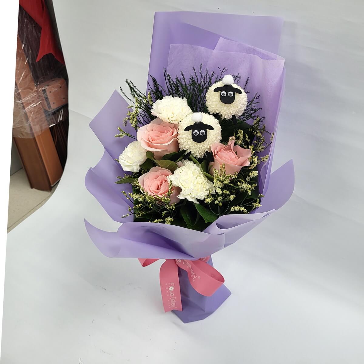 Charming bouquet with pink roses, white carnations, and two adorable sheep-shaped pompom chrysanthemums, wrapped in lavender paper with a pink ribbon. 可愛羊咩咩花束，甜美粉色與柔白色調，充滿童趣與溫馨氣息。