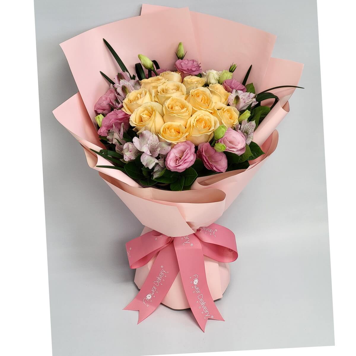 Soft yellow roses surrounded by lilac eustoma and alstroemeria, wrapped in pastel pink paper with a matching pink ribbon. 柔黃玫瑰與粉紫桔梗和六出花交織，淡粉包裝營造溫柔甜美氣息。