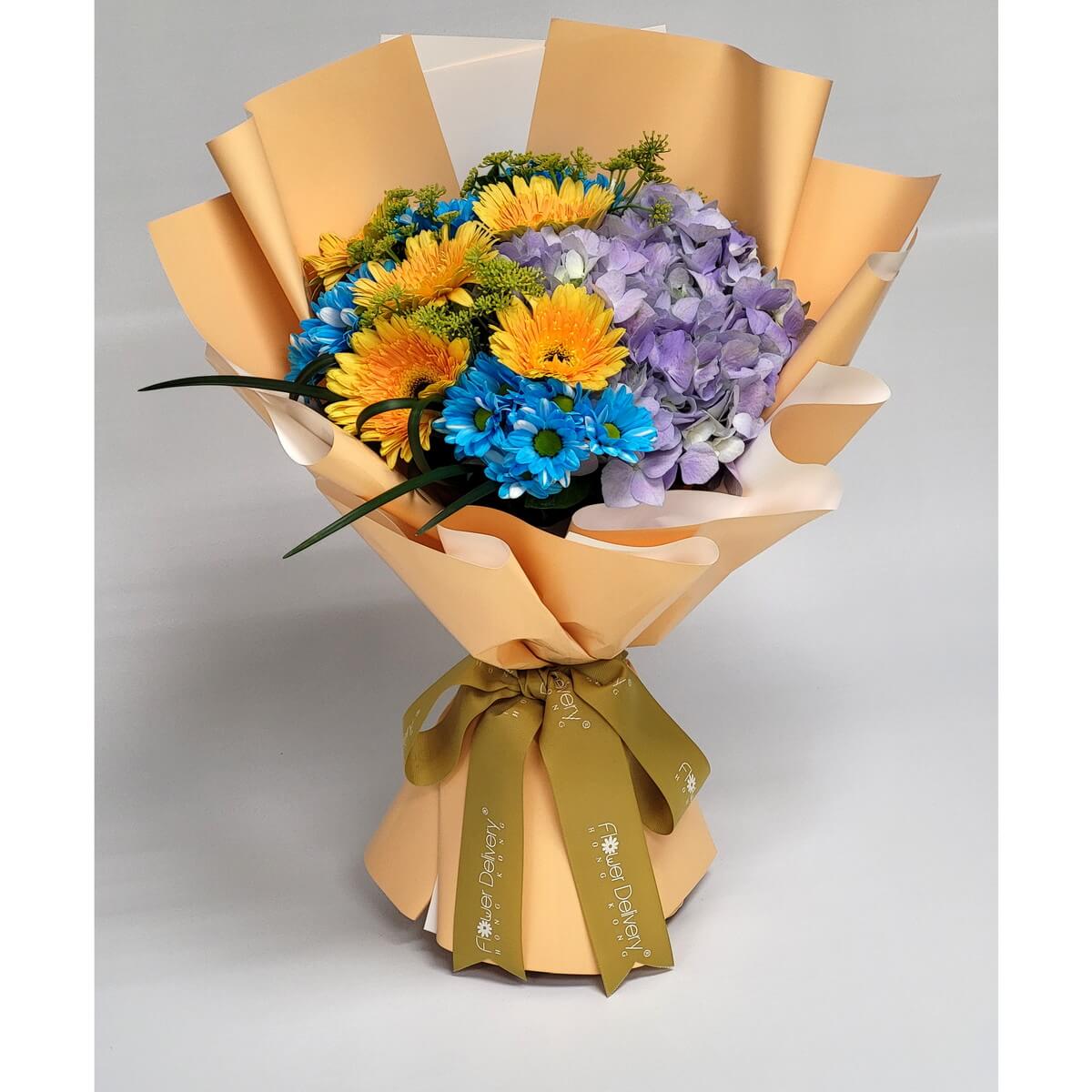 Bright yellow, blue, and violet blooms featuring gerberas, chrysanthemums, and hydrangeas, wrapped in warm apricot paper with a golden olive ribbon. 色彩繽紛的太陽花與繡球花搭配杏色包裝與橄欖綠絲帶，洋溢朝氣。