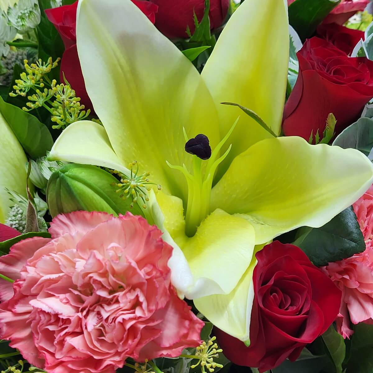 Large yellow lily at center, accented by red roses, pink carnations, green buds, and foliage. | 中央黃百合搭配紅玫瑰、粉紅康乃馨、綠色花苞及豐富綠葉。