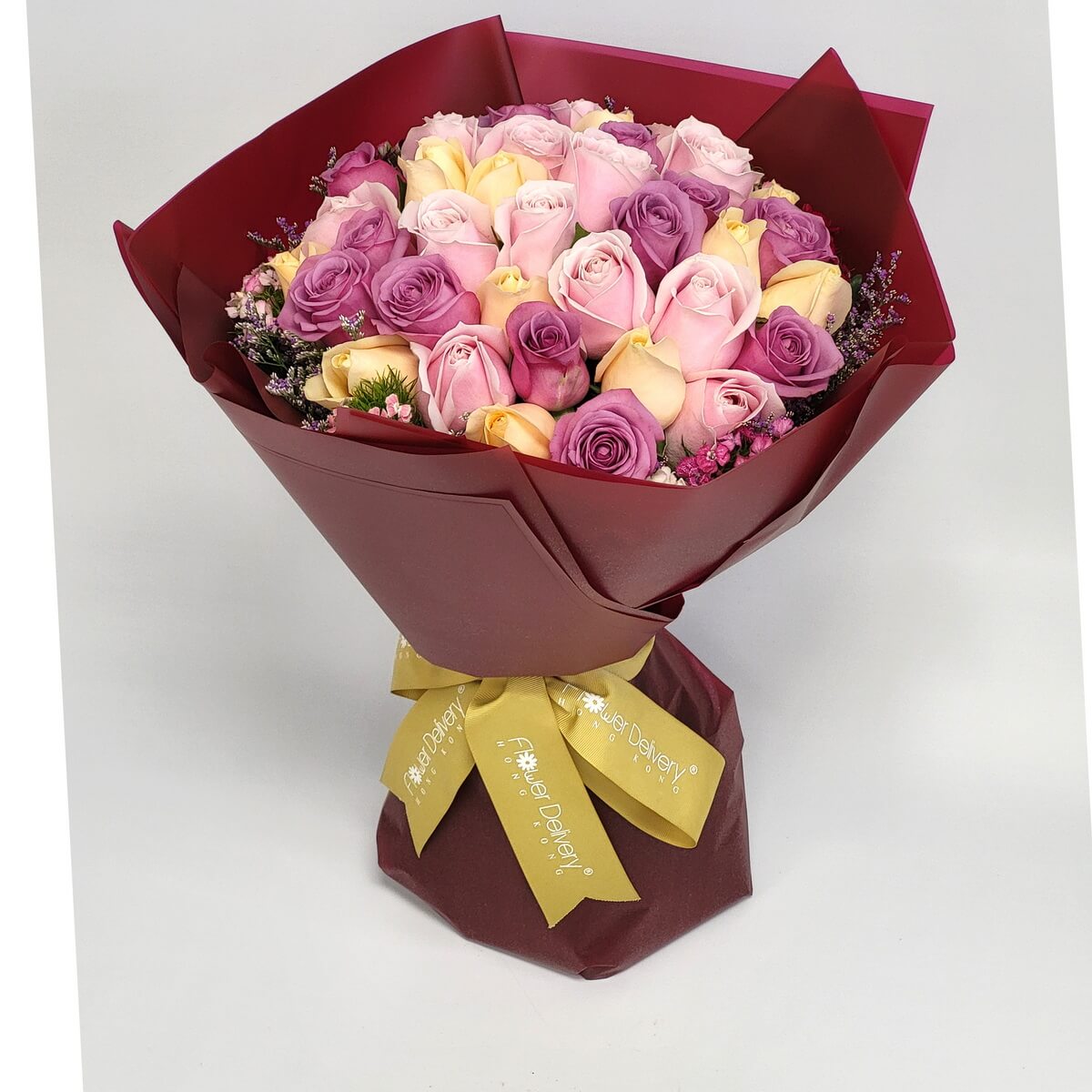 A luxurious bouquet of pink, lavender, and champagne roses arranged tightly in a dome shape, accented with heather and greens, wrapped in deep red paper with a gold ribbon. 粉紅、薰衣草紫與香檳玫瑰交織成圓頂花束，搭配紅色包裝紙與金緞帶，典雅迷人。