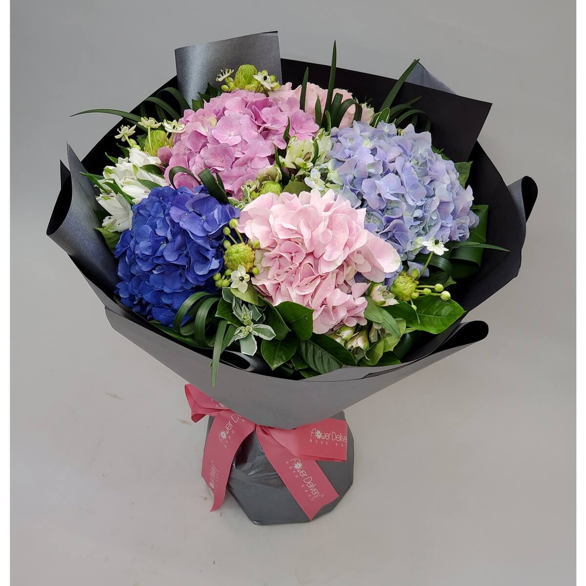 Bouquet with pink, blue, and lavender hydrangeas, white flowers, green buds, and foliage in black wrapping with a pink bow – 粉、藍、紫繡球花配白花與綠葉，黑色包裝，粉紅絲帶點綴