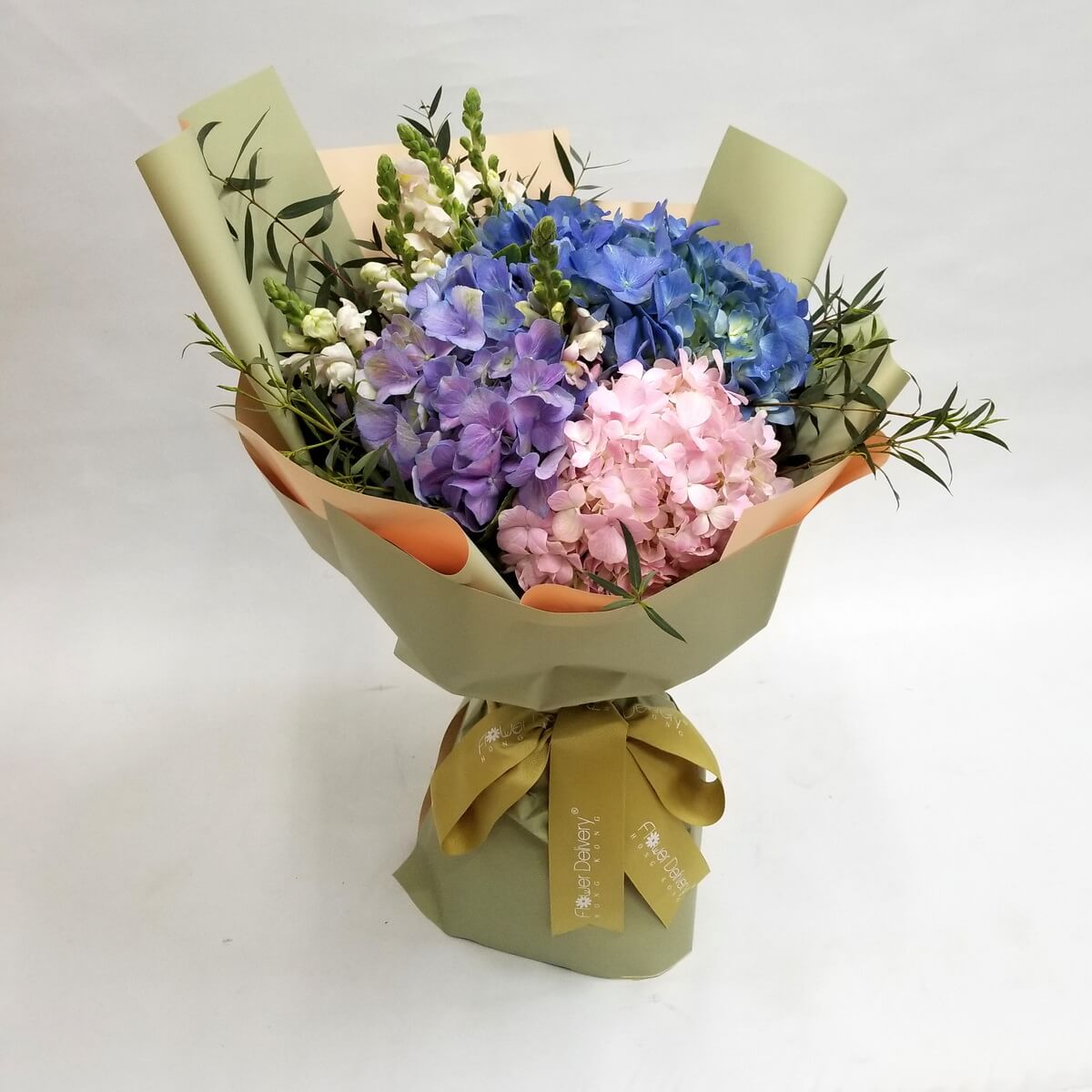 A delicate bouquet of blue, purple, and pink hydrangeas accented with white snapdragons and greenery, wrapped in olive green and peach paper with a mustard yellow ribbon. 柔和色系繡球花與龍頭花，綠橄欖包裝清新自然。