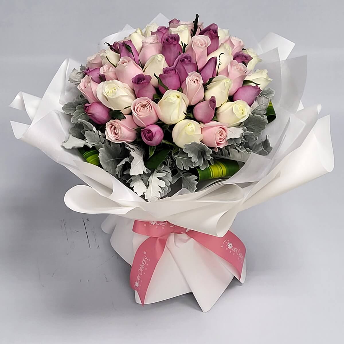 Bouquet of ivory, blush pink, and lavender roses, framed with silver dusty miller leaves and wrapped in white paper with a soft pink ribbon. 淡紫、粉紅與象牙白玫瑰，搭配銀灰葉材與白色包裝，甜美高雅。
