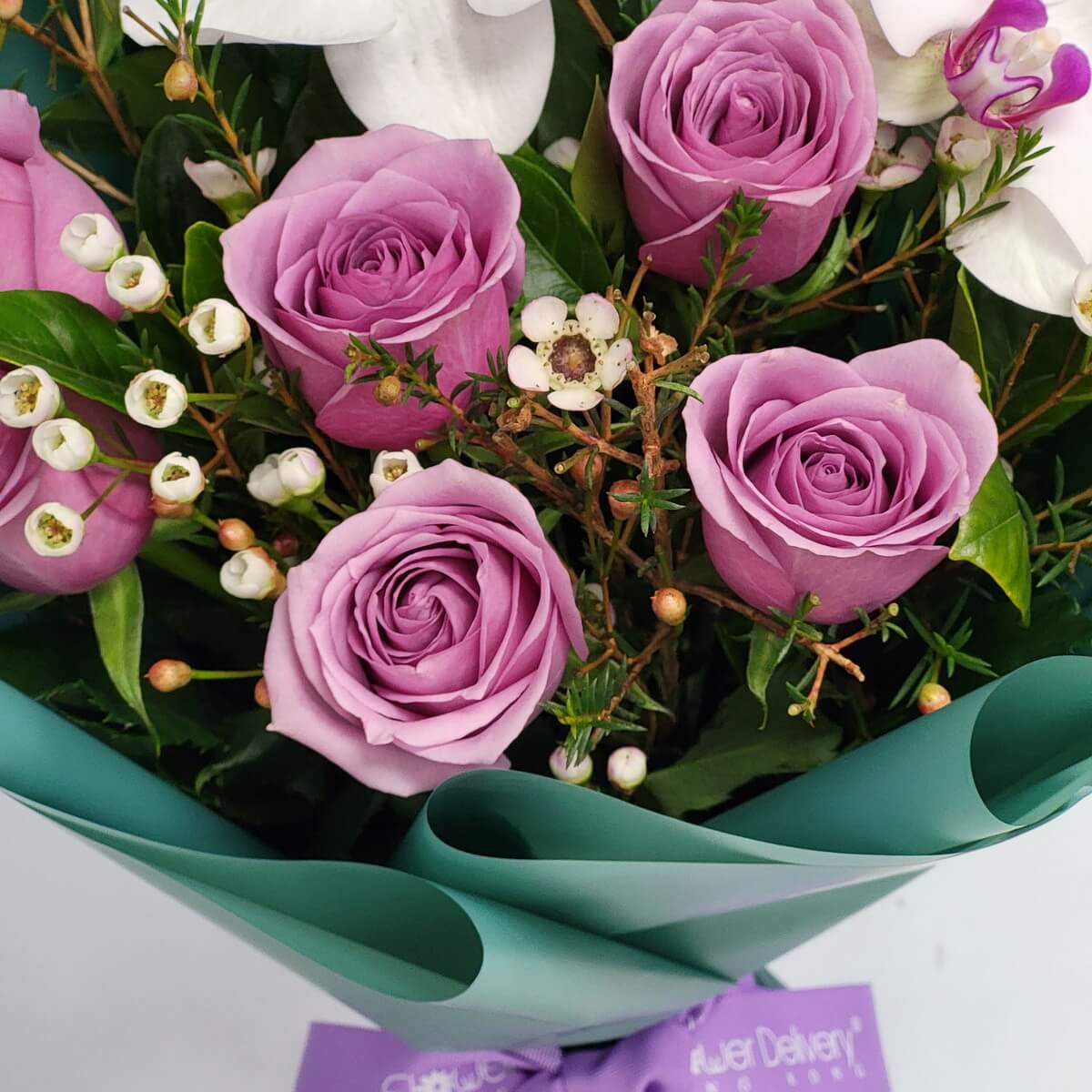 Bouquet featuring lavender roses, delicate white waxflowers, and lush greenery, wrapped in glossy teal paper – 淺紫玫瑰與白色蠟花、青綠葉混搭，包裹於亮面青綠色包裝紙中