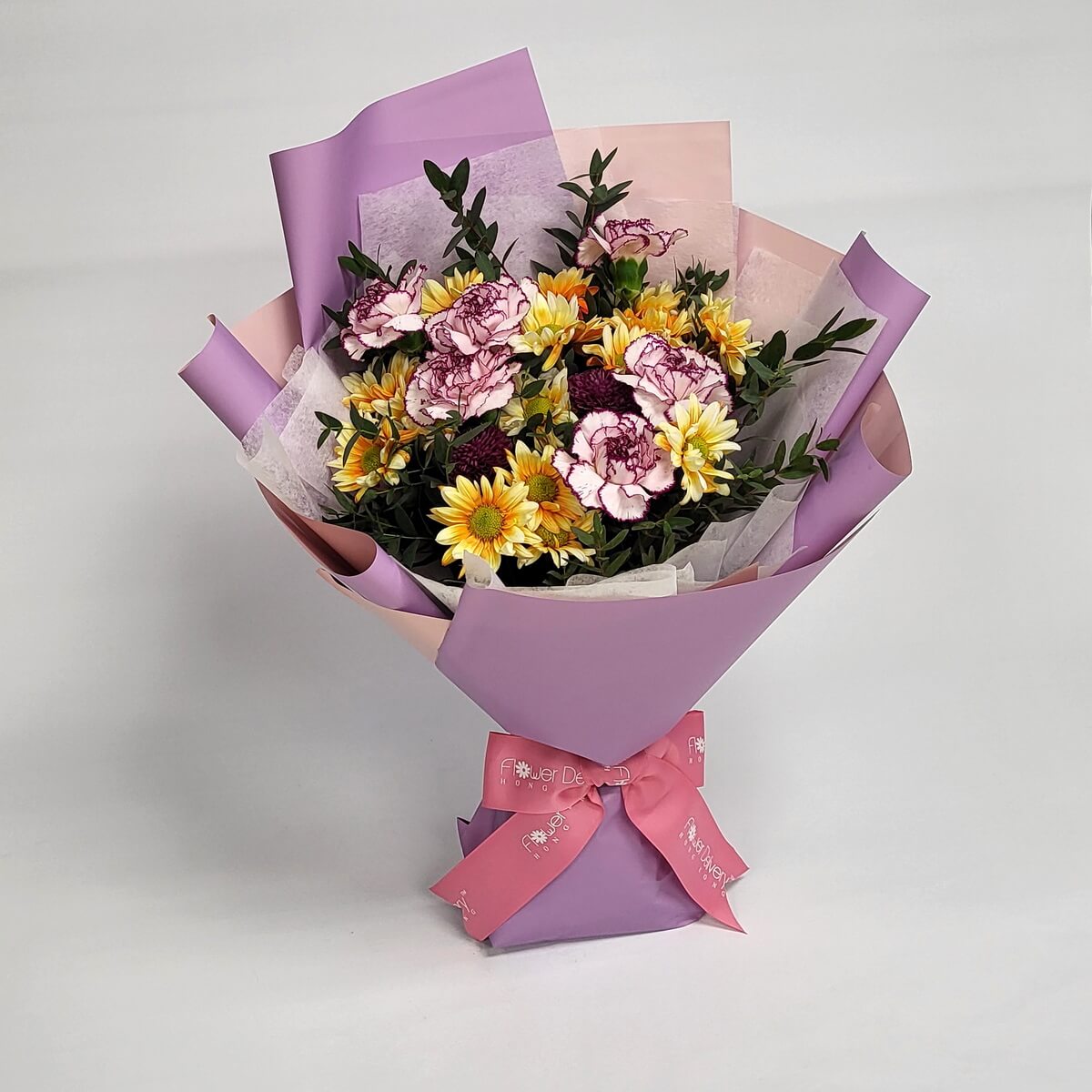 Yellow daisies and purple-edged carnations with greens, wrapped in lavender and peach-toned paper with a pink ribbon. Fresh and joyful. 黃雛菊與紫邊康乃馨搭配嫩綠葉材，薰衣草與蜜桃包裝，展現清新快樂氣息。