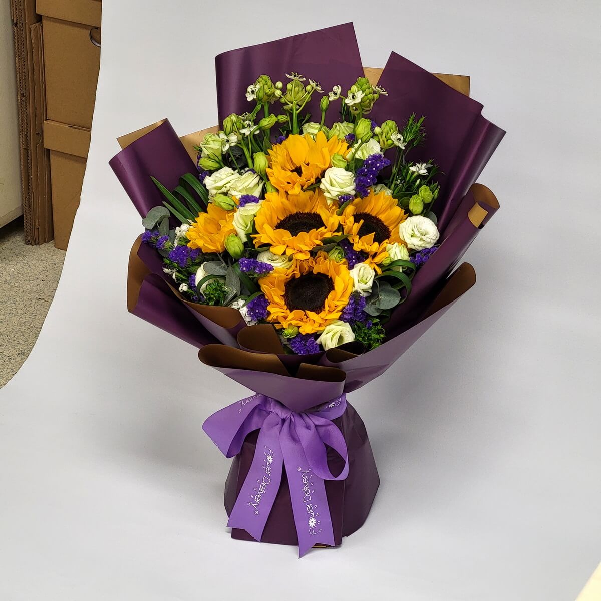 Bright sunflowers with purple statice, white eustoma, and lush greenery, wrapped in rich purple and gold paper with a lavender ribbon. Sunny and cheerful. 向日葵配紫色勿忘我與白桔梗，紫金包裝，洋溢陽光氣息。