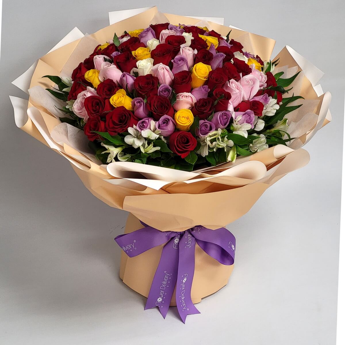 Large bouquet of multicolored roses—red, pink, lavender, yellow—wrapped in champagne and white paper with a purple ribbon. 色彩繽紛的大型玫瑰花束，紅粉紫黃交織，配香檳白包裝與紫色絲帶，活潑華麗。
