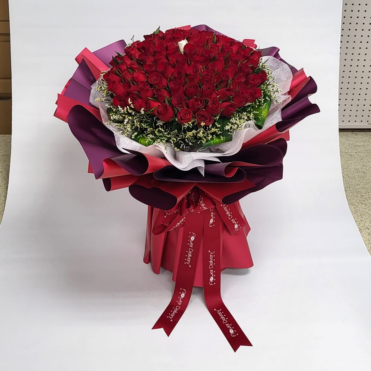 Stunning large bouquet of red roses with a single white rose at the center, surrounded by fillers and wrapped in layered red and purple paper. 巨型紅玫瑰花束，中央一朵白玫瑰，配襯滿天星與紅紫包裝紙。